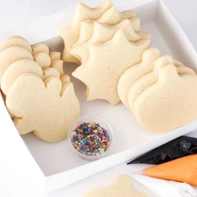 A white box containing decorated sugar cookies in holiday shapes, including a mouse, snowman, Christmas tree, and pumpkin, along with a small container of rainbow sprinkles, a black piping bag, and an orange piping bag.